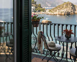 A panama hat lies discarded on a private balcony chair. The view stretches across the Bay of Mazzarò to a white rocky outcrop