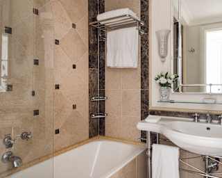 Elegant bathroom with beige tiled shower and bath, glass divider, white basin, large mirror, wall shelf with towels, chrome fittings, and a small vase with white flowers on the worktop.