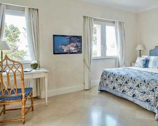 A marble floor and blue accents give this bedroom a cool feel. Tall trees can be seen in bright sun through two large windows