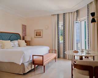 A Deluxe Sea View Suite with a white bed, coral bench, and a marble table with two chairs by glazed doors with long drapes.