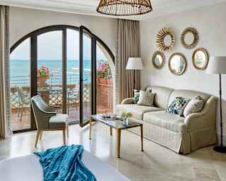 Small boats with canopies bob on the sea. Arched glass doors give access to the private balcony from an elegant sitting room