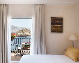 Across a king bed made with impossibly crisp white sheets, the Bay of Mazzaro headland stands proud of the deep blue sea