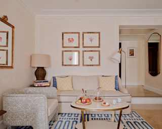 A table of fruit and cake sits on a blue and white rug flanked by a grey chair and pale sofa, with four red wall prints above.