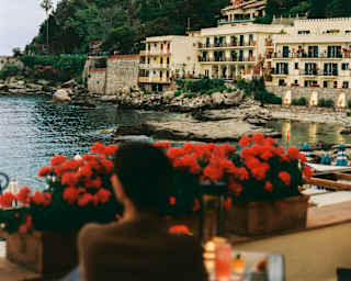 A guest in soft-focus sits at bar table overlooking Mazzarò beach, with a view of the hotel at one end, seen from behind.