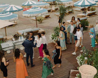 Well-dressed guests mingle on a deck behind the parasols and loungers on Mazzarò beach, seen from above at an evening event.