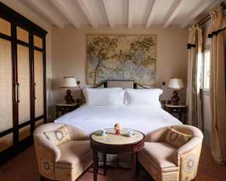 In a serene room, two peach tub chairs and a wooden table sit at the end of a white bed with a tapestry on the wall behind.