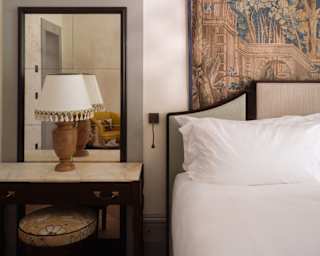 View of a bedside space, with a round wall mirror above a rectangular one propped on a side table with a reading lamp by a bed.