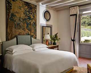 A detailed woodland tapestry hangs above a double bed with white linen in a Premium Room with large windows on to gardens.