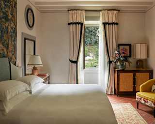 View across the white linen of a bed to a window, wide open on to gardens, with a vintage cabinet and chair to the right.