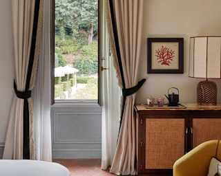 A large window with tied-back drapes opens on to lush gardens, seen from the end of a bed with a glimpses of furniture.