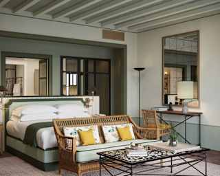 A wicker sofa with yellow and floral cushions abuts a white bed with hints of mint, seen over a low table in a Garden Suite.