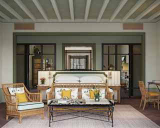 A table with a wicker chair and sofa abuts a bed, in a front view of a Garden Suite with wall plates, desk and green décor.