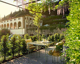 A patio table for two, with tea, sits under a sun-dappled wisteria-clad pergola with views of the hotel and verdant grounds.