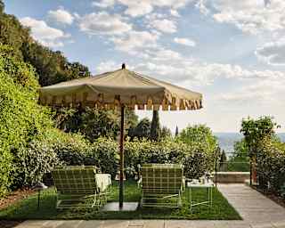 Two green-cushioned loungers recline under a parasol on a lawn terrace on a hillside with wonderful views, seen from behind.