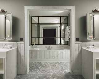 View through a wall arch, flanked by basin units, to bath area with a mirrored wall, seen in a marbled Junior Suite bathroom.
