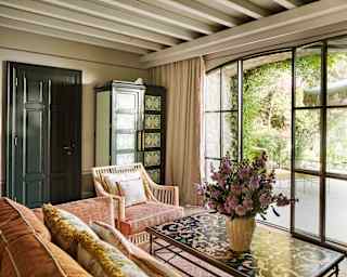 A bright living area with coral sofa, cane chair, floral inlay table with a vase of pink blooms, and glazed terrace doors.