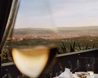 A close up image of a glass of white wine fades into a long view of Florence, its white walls and red roofs surrounded by green