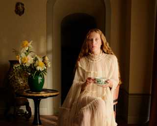 A woman in a long, sheer, light dress sits barefoot on a chair indoors, holding a teacup. She sits beside a small table with a vase of yellow flowers, in a softly lit room with an arched doorway behind her.