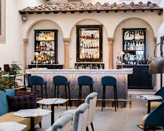 View of a lounge with grey chairs, hexagonal marble tables and a bar, with four stools and bottle shelves in arched alcoves.