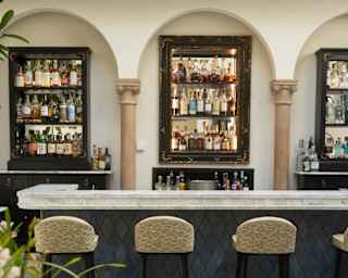 Four stools line a white marble bar with teal front facing three gleaming bottle shelves spaced between decorative wall arches.