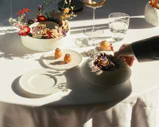 Three dishes of amuse-bouches, two decorated with bright flowers, sit on a white-clothed table in half-sun with a glass of wine.