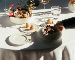 A sunlit table set for two with elegant plates, floral centrepiece, wine glasses, and a hand reaching for a bowl of food. A person is seated in the background, partially in shadow.