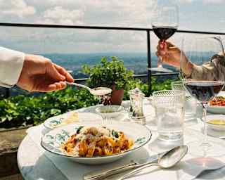 A waiter sprinkles parmesan onto an unctuous pasta dish as a guest opposite raises a glass of red wine, at an al fresco table.
