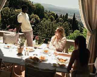 Tilted image of a man standing by a table on a covered terrace with tied drapes, where guests enjoy lunch with sublime views.
