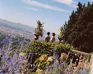 Angled image of two guests standing on the stone terrace in a garden bursting with lavender and foliage overlooking Florence.