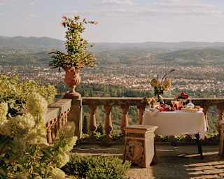 A table piled with food awaits guests on a rustic balustraded terrace overlooking sunny Florence and Tuscan hills beyond.