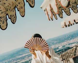 A woman in a diaphanous short white dress holds a fan in front her face as she stands in sunny gardens between parasols.