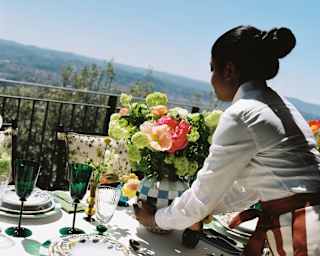 A female staff member places a vase of green and pink ranunculi on a sunny, view-filled terrace lunch table, seen from behind.