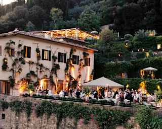 Guests mingle on the terrace as warm window and garden lights provide a magical evening atmosphere, seen across a courtyard.