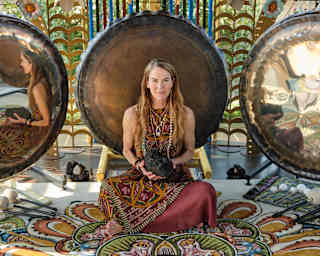 J.J. Martin, with long blonde hair and ethnic dress, sits in a sanctuary space in front of three large gongs holding a lava rock.
