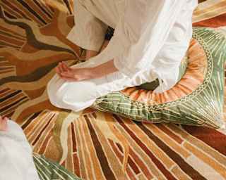 A woman in white, seen from the shoulders down, sits in meditation pose on a cushion and throw in browns and greens.