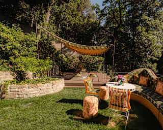 View of a canopied deck behind a lawn with a curved La DoubleJ floral fabric sofa, table, two cushioned chairs and stools.
