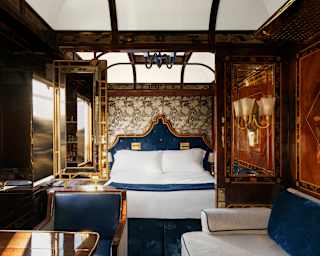 View through Venice Suite's living area and gilt-framed mirror partitions to a bedroom with white bedding and navy headboard.