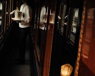 A steward in white jacket stands half in shadow in a carriage corridor holding a tray with a wine cooler and bottle of champagne.