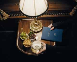 A guest's hands hold a blue book titled Writers on the Rails, and a drink, seen from above at a lamp-lit table with snacks.