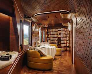 A lounge view glances a window-side daybed with book and binoculars into a calm space with yellow chairs, table and library.