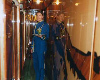A smiling female train attendant in a blue uniform stands in a narrow, glossy train corridor, holding a drink on a tray.