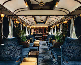 A view through the decadent old-world Bar Car with banquettes and carpet in midnight blue with gold stools and glowing lamps.