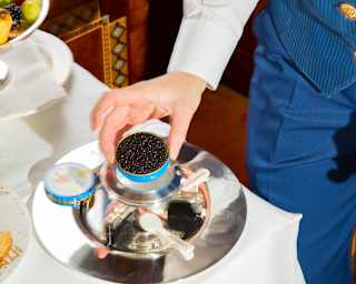 A steward in a blue waistcoat and trousers, seen from the shoulders down, sets a tin of caviar onto a silver tray on a table.