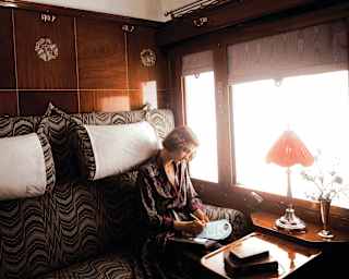 In an Art Deco vision, a woman with a blond bob in a silk gown writes a letter in the glow of daylight by her cabin window.