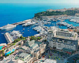 Aerial view of a marina filled with yachts and boats, surrounded by modern and historic buildings, with the sea and hills in the background.