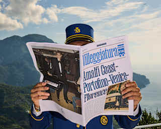 A man in a blue train steward's uniform holds open a Villeggiatura newspaper, obscuring his face, on an Amalfi-view terrace.