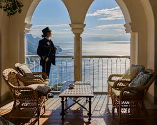 A woman in a train steward's uniform stands in the left space of a double arch of an open-sided sunroom overlooking the sea.