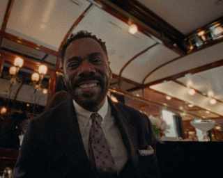 Colman Domingo, in a jacket, shirt and tie, smiles to camera in a low-angle image capturing interior carriage lights above.