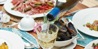 White wine is poured into a crystal glass as guests enjoy a feast, with plates of charcuterie, lentils, salad and shellfish.