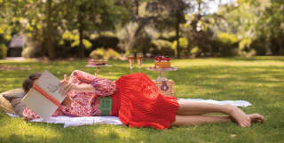 A young woman lies on the grass reading a book; behind her is a sumptuous picnic of fruit topped cakes and sparkling rosé